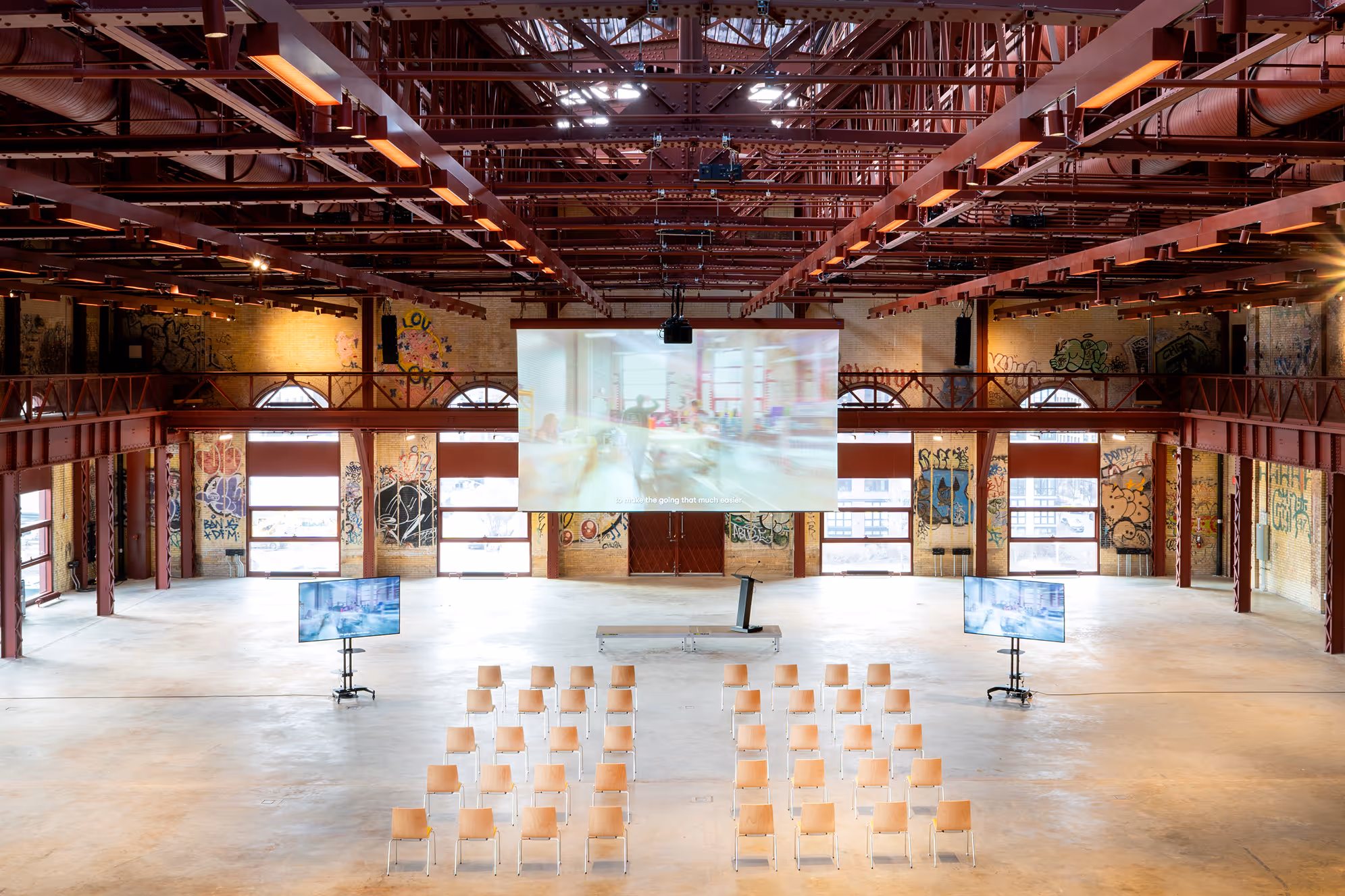 Industrial-style event hall with rows of wooden chairs facing a podium and large projection screen, surrounded by graffiti on brick walls.