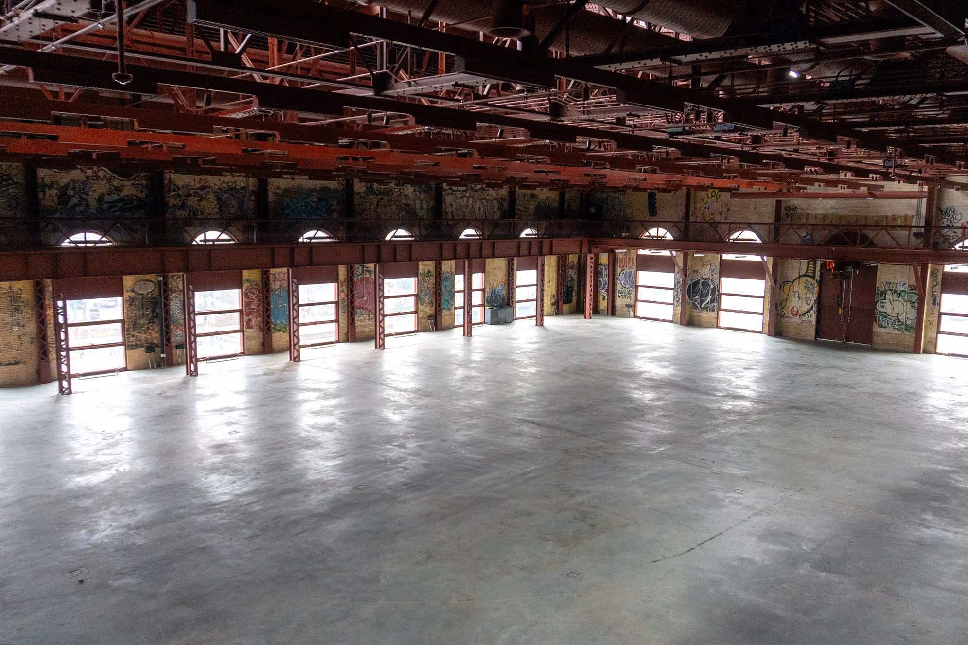 Large empty industrial warehouse with concrete floor, graffiti-covered walls, red metal beams, and multiple windows.
