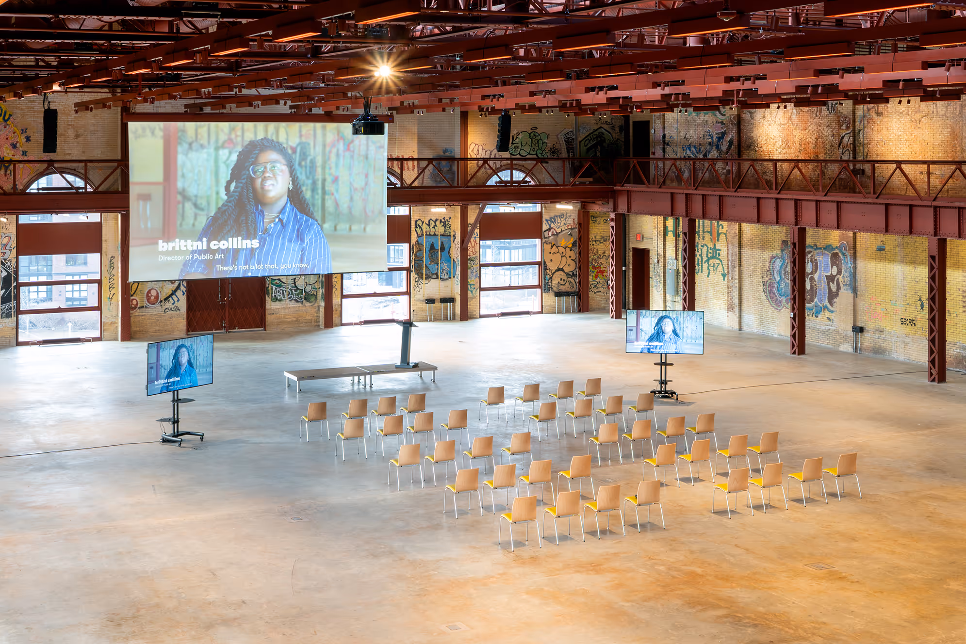 Large industrial-style hall with exposed red beams, graffiti walls, and a seating arrangement facing a stage with three screens showing a woman identified as Brittni Collins, Director of Public Art.
