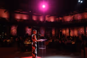 Woman in a floral dress speaking at a transparent podium in a dimly lit event space with purple spotlight and seated audience.