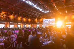 Guests seated at tables in a warmly lit banquet hall with a large screen and bright sunlight streaming through windows.