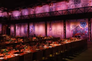Long banquet table set with floral centerpieces and candles in a dimly lit industrial space with purple graffiti-covered walls.