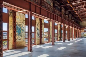 Interior of an abandoned industrial building with rusted red steel beams and graffiti-covered walls, illuminated by natural light through tall windows.