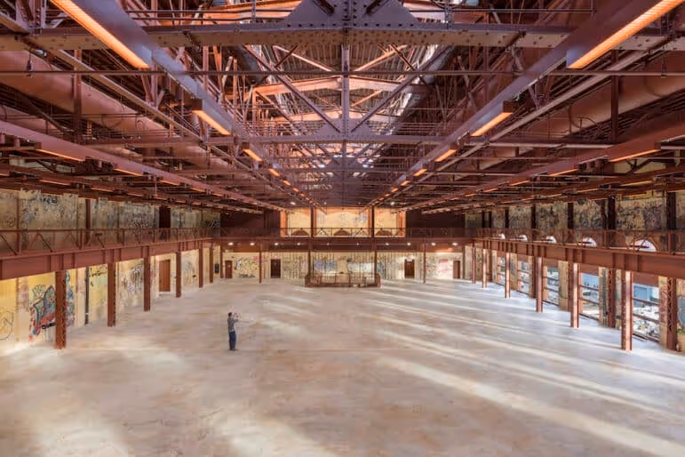 Spacious empty industrial warehouse with exposed rust-colored steel beams and a person standing in the center.