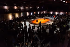 Large group of people gathered around a circular stage with orange lighting in a dimly lit venue.