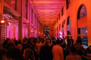 Crowded indoor event space with high ceilings and large arched windows, illuminated by pink and red lights.