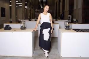 Woman in a white sleeveless top and black-and-white skirt standing between two white display tables in a spacious room.