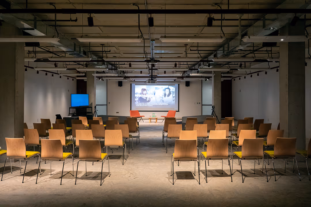 Empty conference room with rows of wooden chairs facing a stage with two orange chairs and a screen displaying a presentation.
