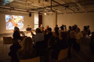 Audience seated in a dimly lit room watching a presentation with a projected image on the wall and two speakers seated at the front.