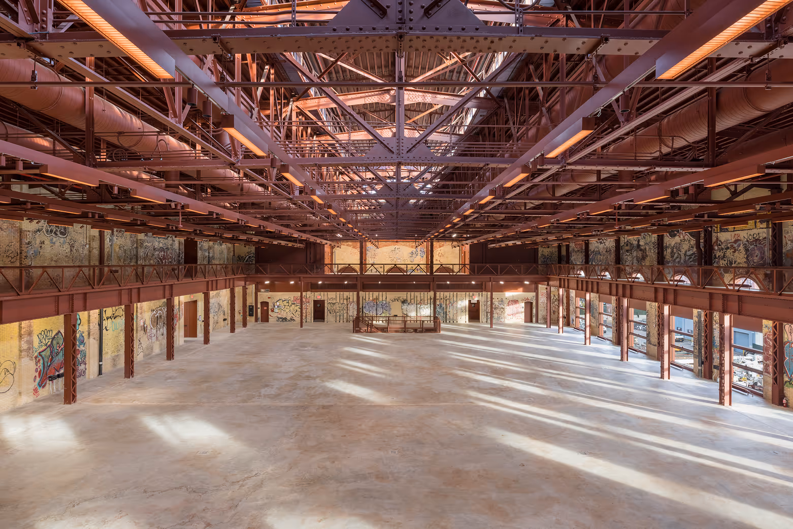 Large industrial-style hall with exposed red steel beams, graffiti-covered yellow brick walls, and empty concrete floor illuminated by natural light through tall windows.