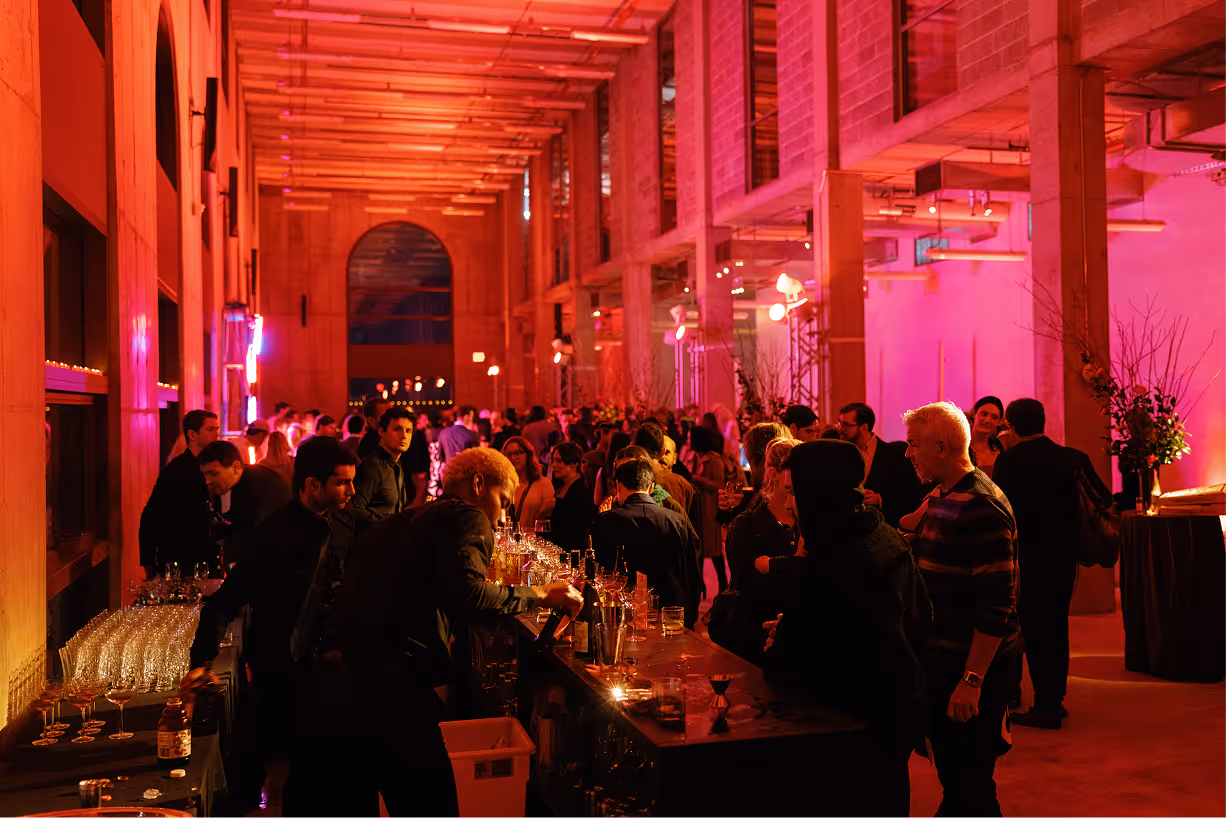 Crowded bar inside a modern venue with warm orange and pink lighting and people socializing.