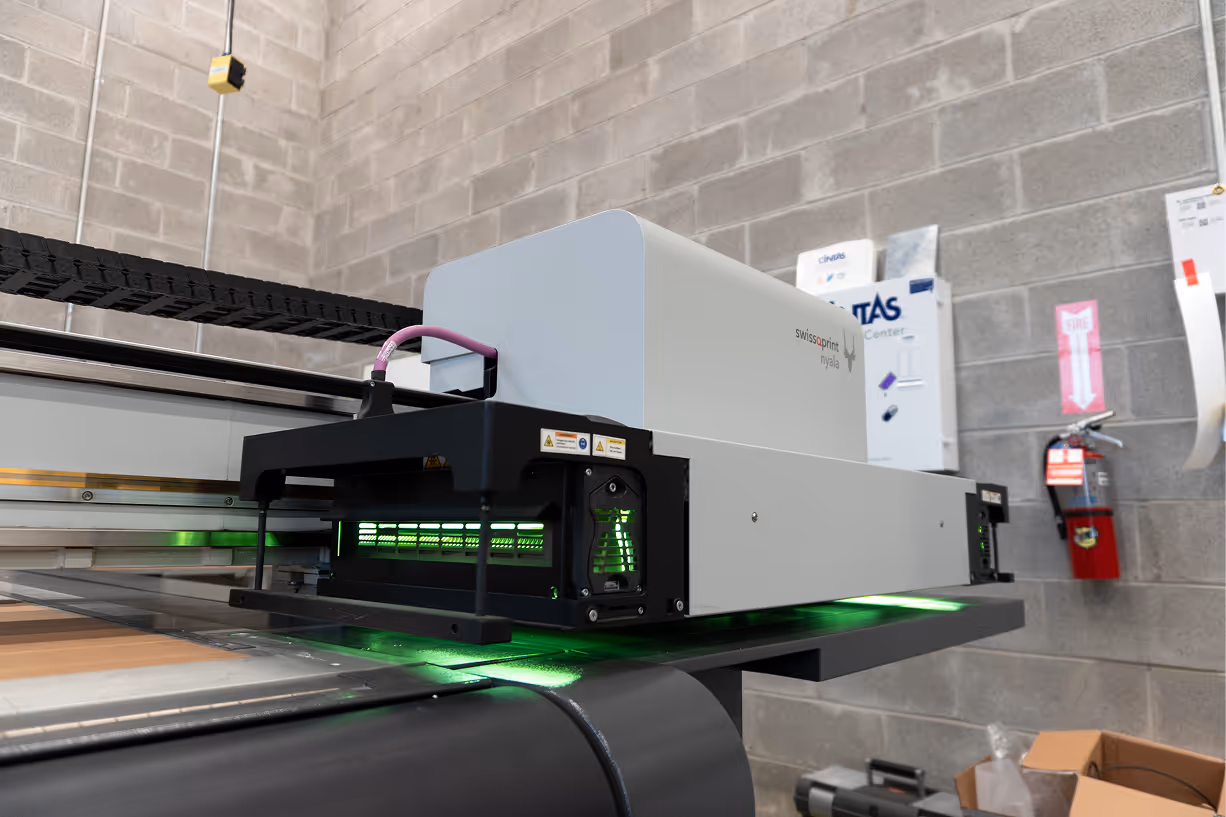 Swiss QPrint Nyala industrial flatbed printer with green light printing indoors against a concrete block wall.