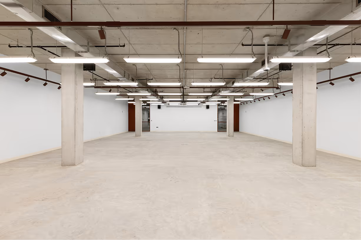 Empty industrial-style room with concrete floor, ceiling, and pillars, illuminated by rows of fluorescent lights.