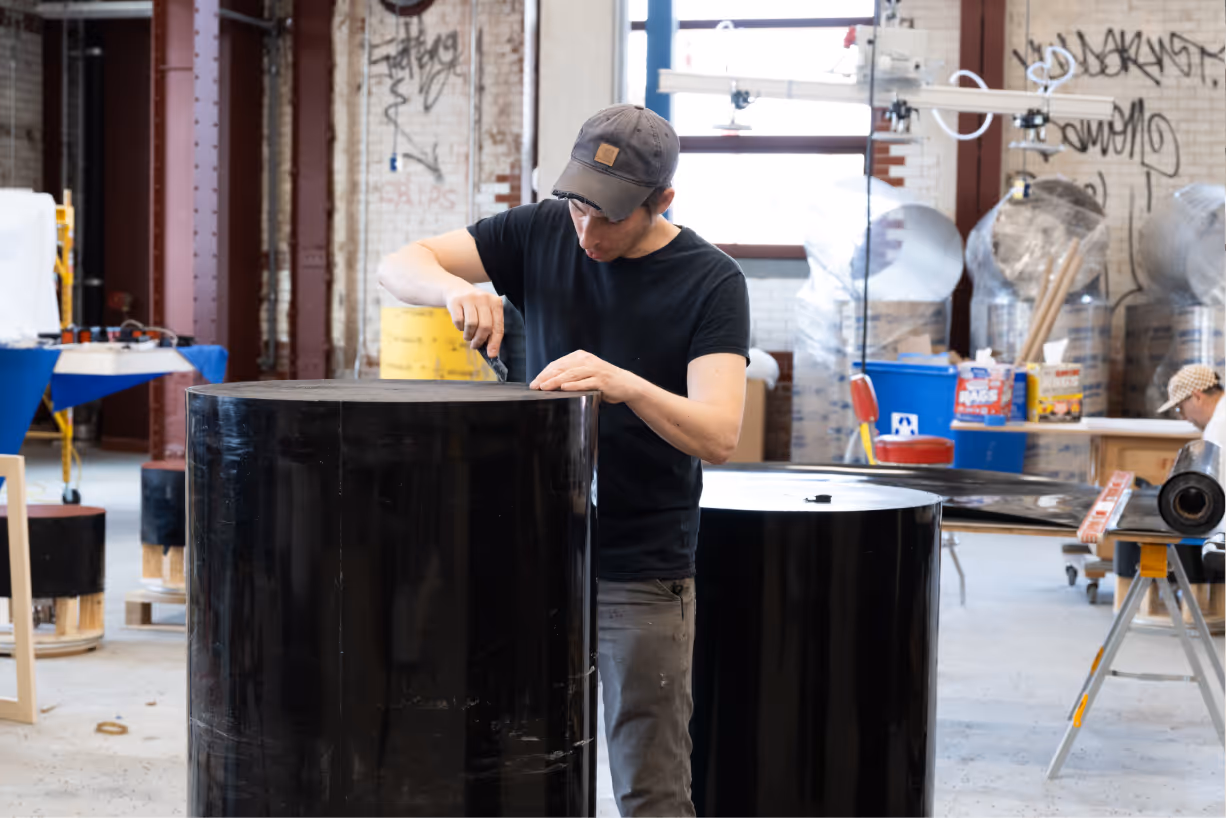 Man wearing a cap scrapes or works on the top surface of a large black cylindrical object in a workshop with industrial equipment.