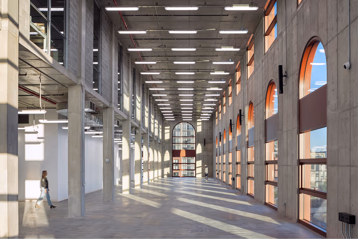 Spacious industrial-style loft with tall arched windows casting long shadows on the concrete floor, featuring two people walking inside.