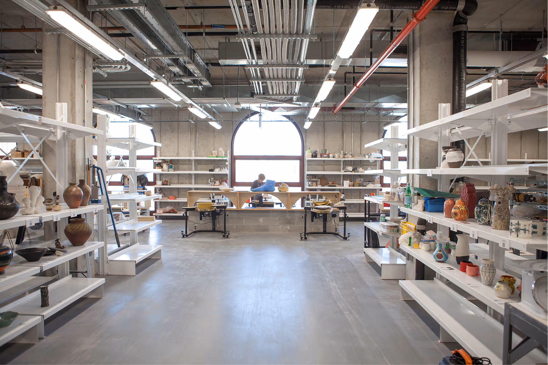 Spacious pottery studio with white shelves displaying ceramic vases and pots, a large arched window, and a person working at a table in the background.