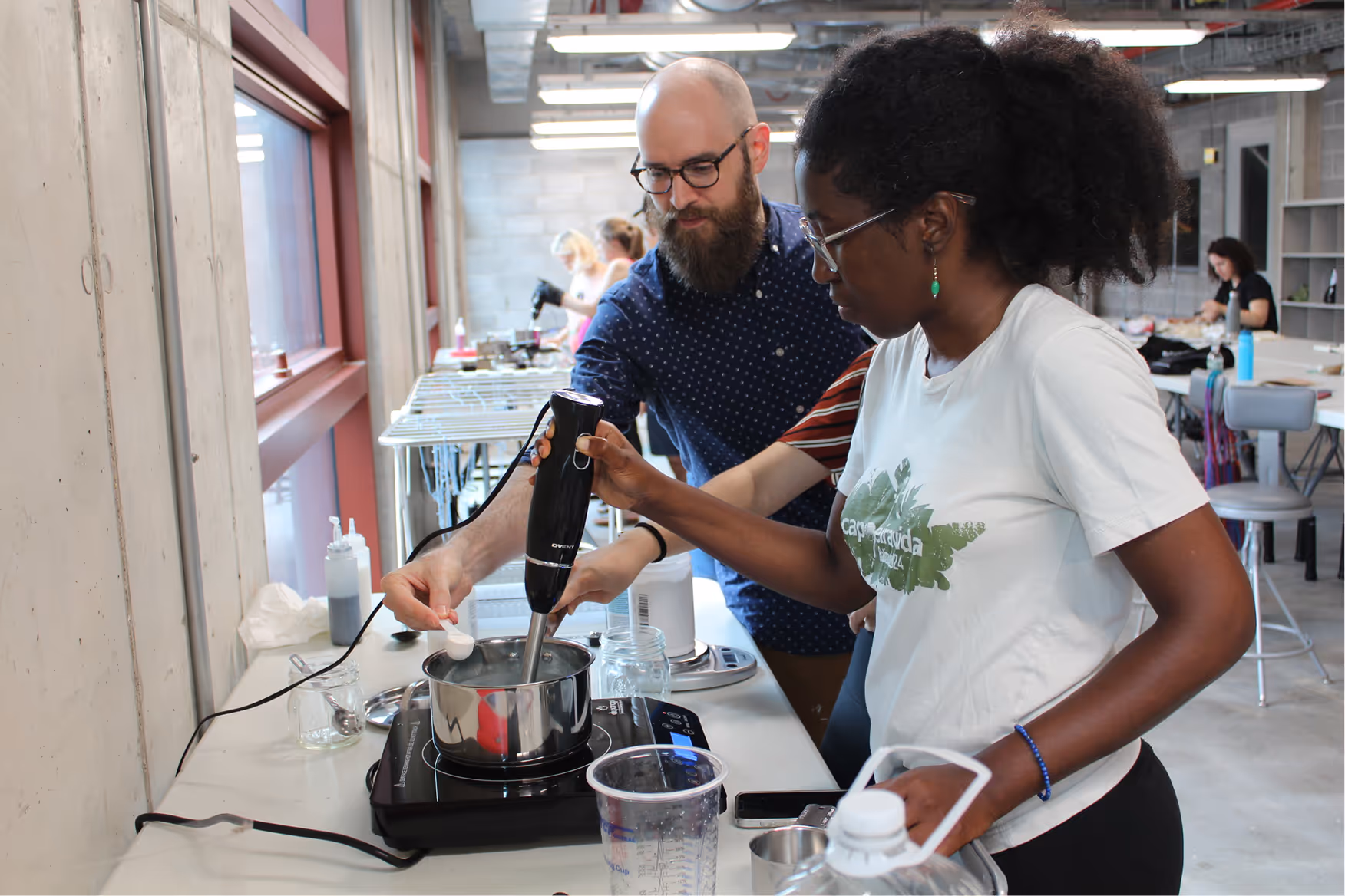 Two people working together to blend ingredients in a pot on an electric stove in a classroom setting.
