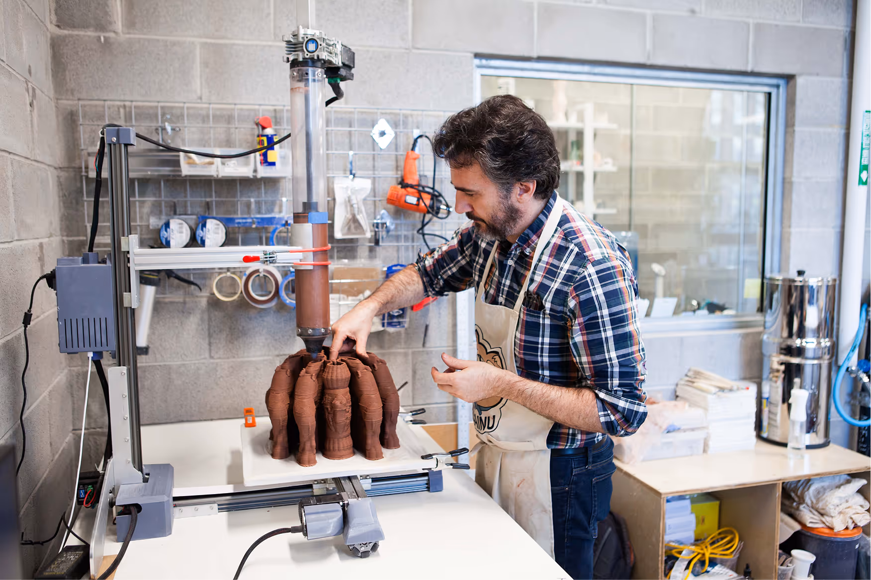 Man in plaid shirt and apron working on a 3D printer creating multiple brown clay sculptures in an art studio.