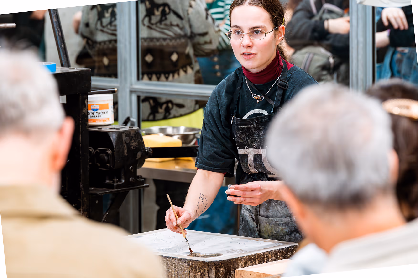 Young woman wearing glasses and apron demonstrating a painting technique to a seated audience.