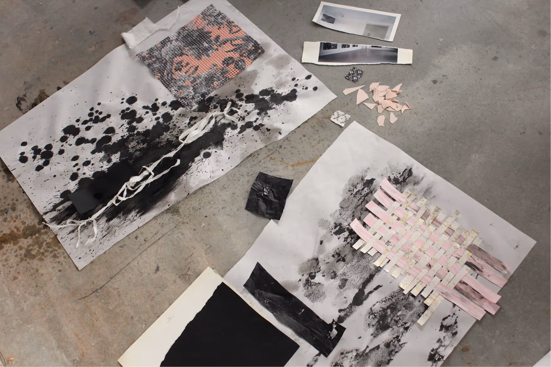 Mixed media art pieces on concrete floor featuring black ink splatters, woven paper strips, fabric, and torn paper elements.