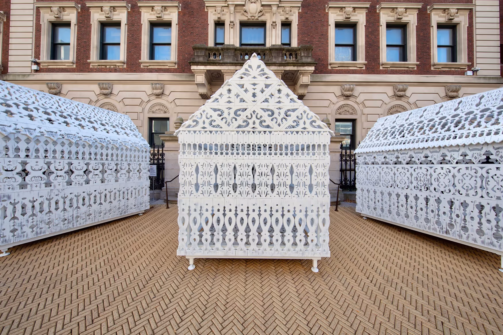 Three white, intricately patterned, lace-like art structures arranged in front of a historic brick and stone building.