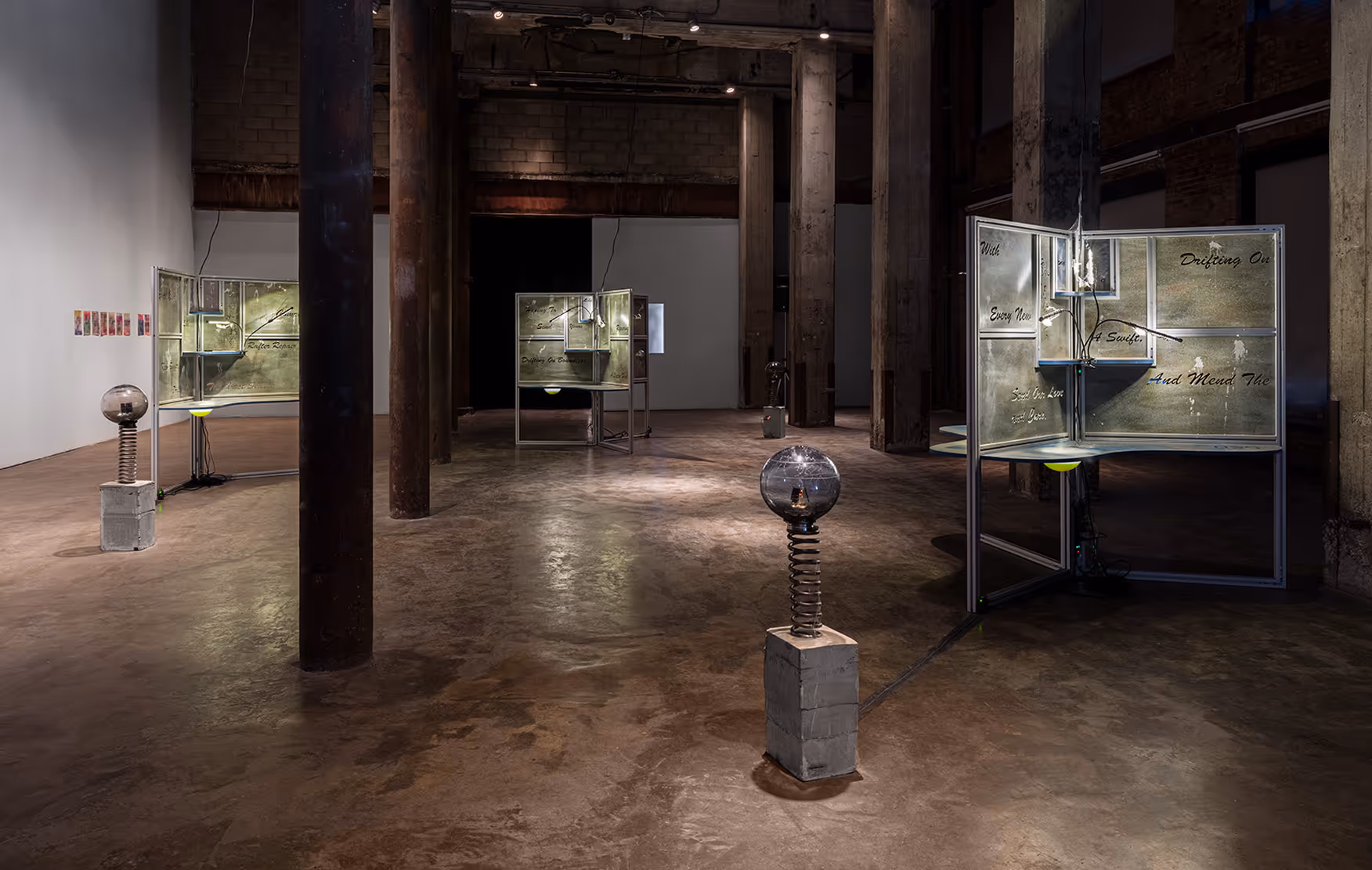 Industrial-style art installation in a large gallery with concrete floors and pillars, featuring illuminated metal panels with handwritten text and spring-supported spherical glass sculptures on concrete bases.