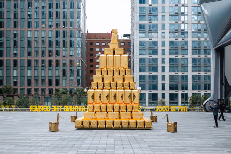 Pyramid-shaped stack of yellow suitcases arranged outdoors between tall modern buildings on a tiled plaza with a few people nearby.