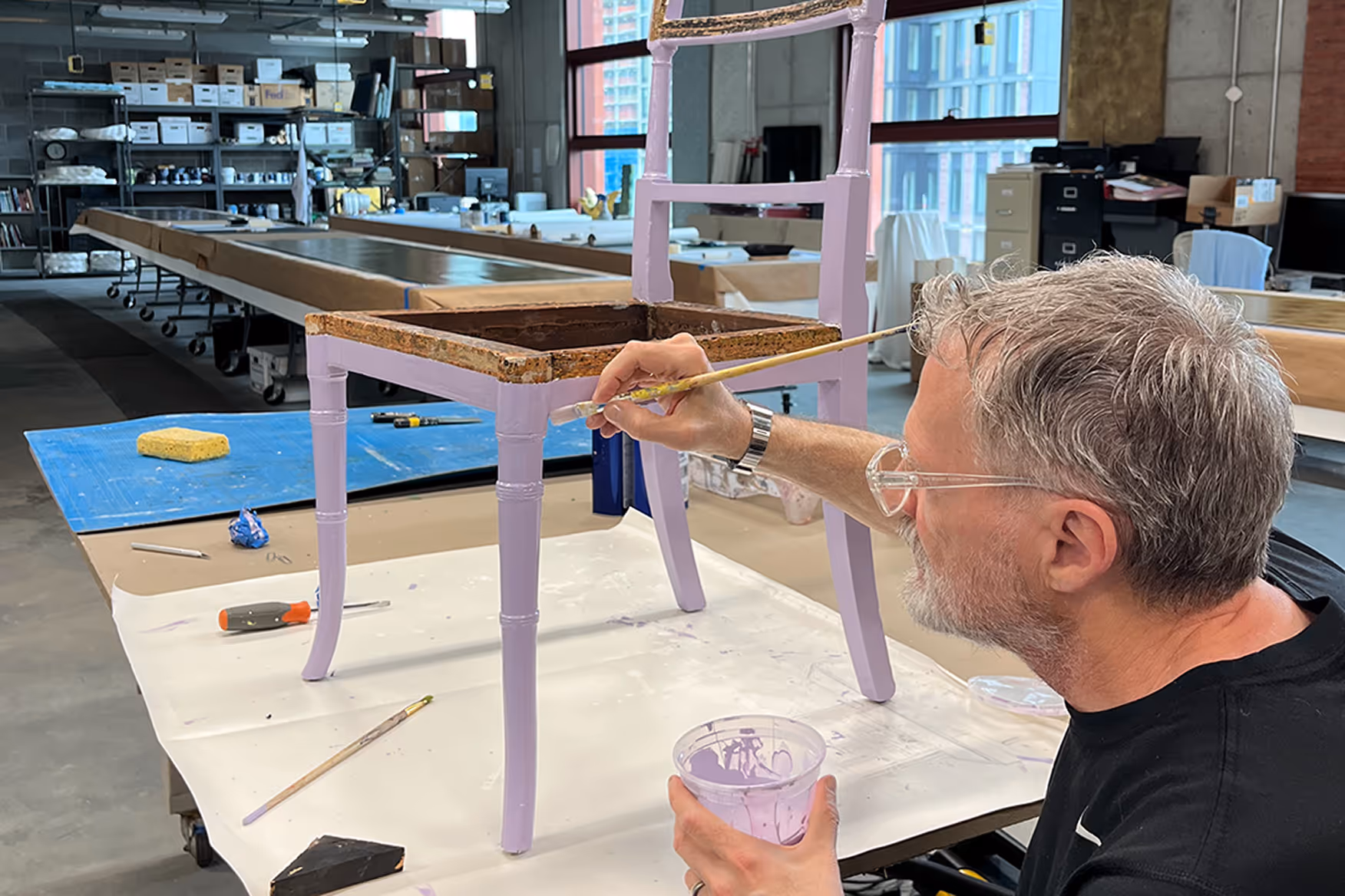 Man with gray hair and glasses painting the legs of a wooden chair lavender in a workshop.
