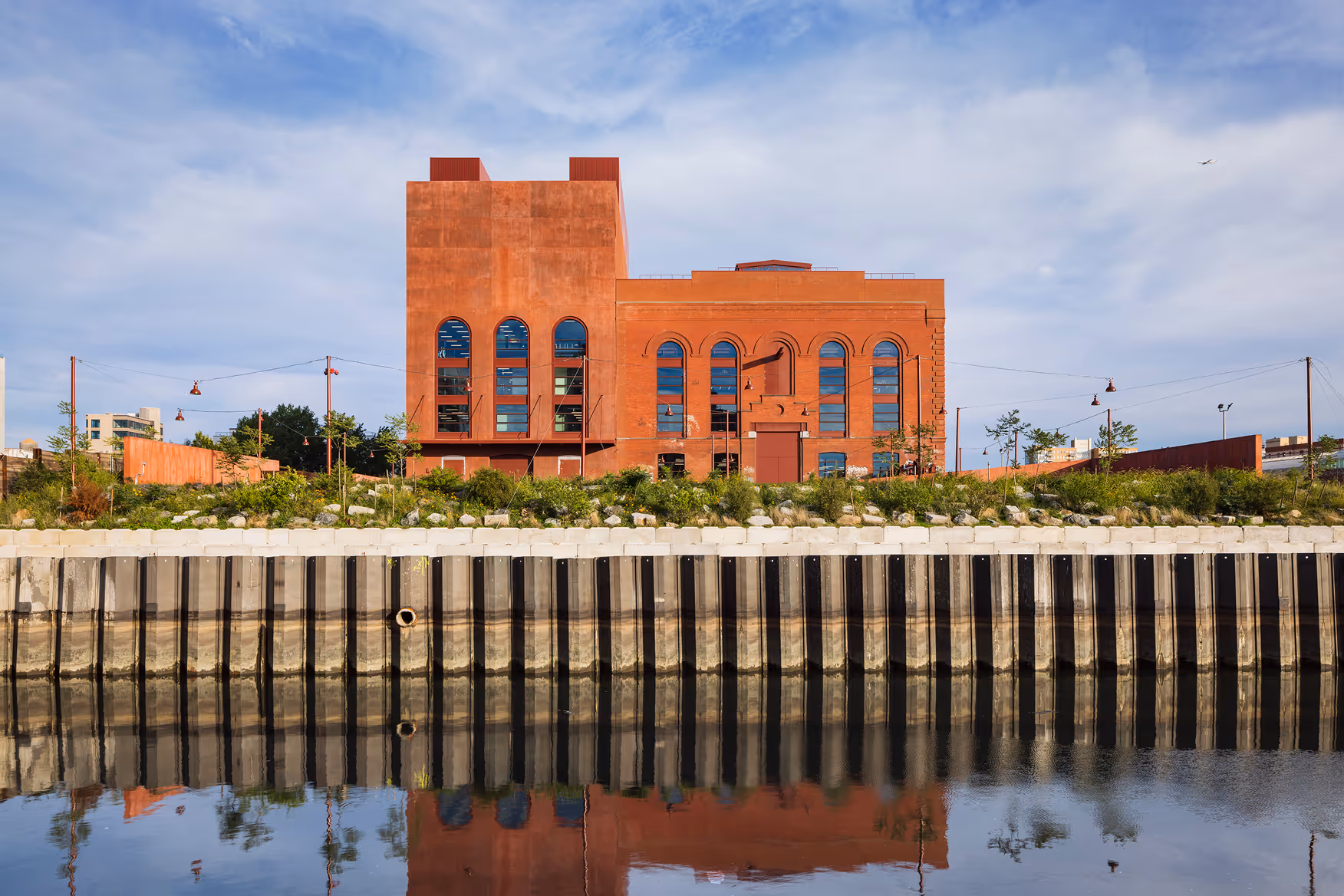 Red brick industrial building with large arched windows reflected in calm river water under a partly cloudy sky.