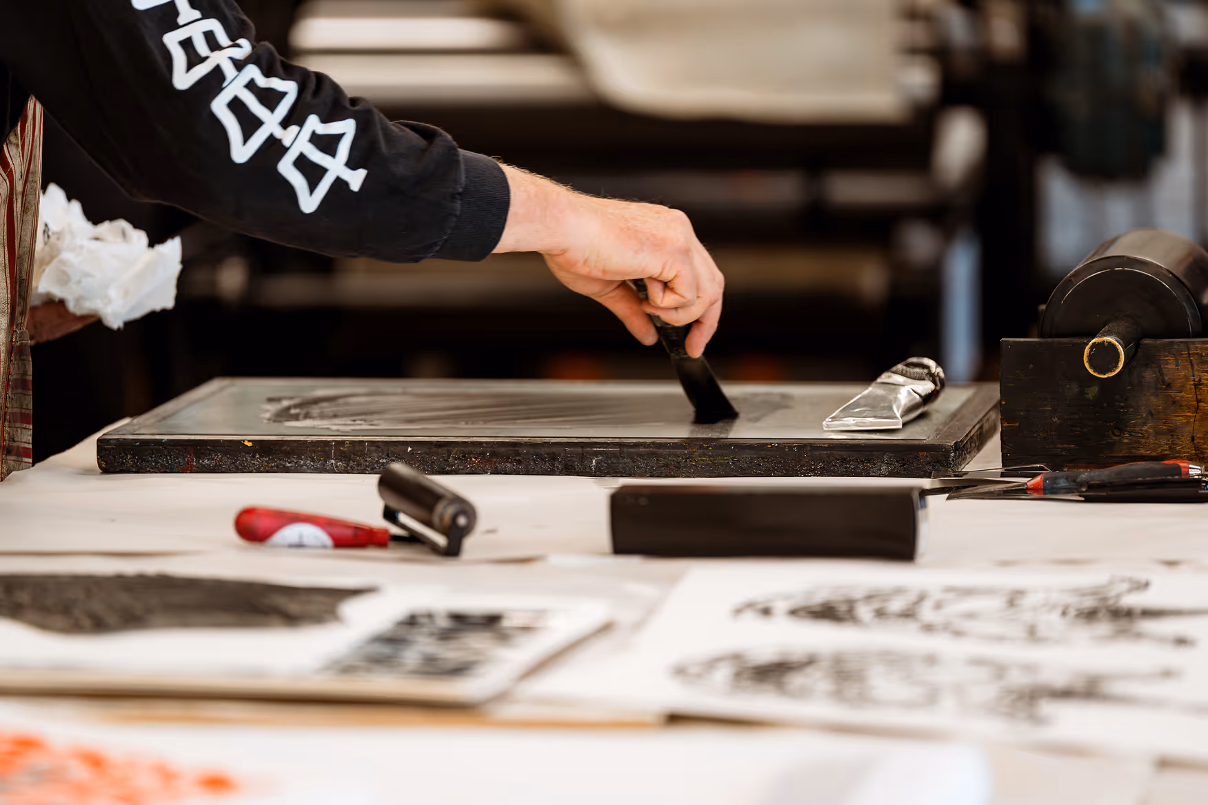 Person using a tool to spread ink on a printing plate in an art studio with printmaking tools and prints on the table.