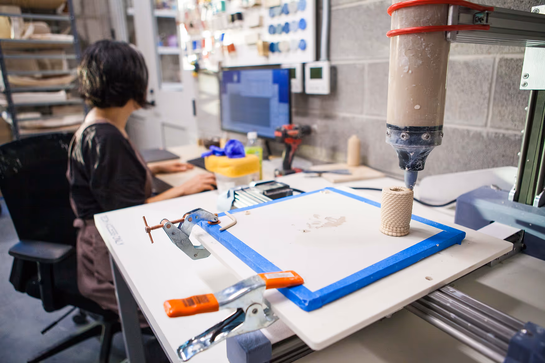 3D printer extruding a textured cylindrical object on a worktable with clamps, a person working on a computer in the background.