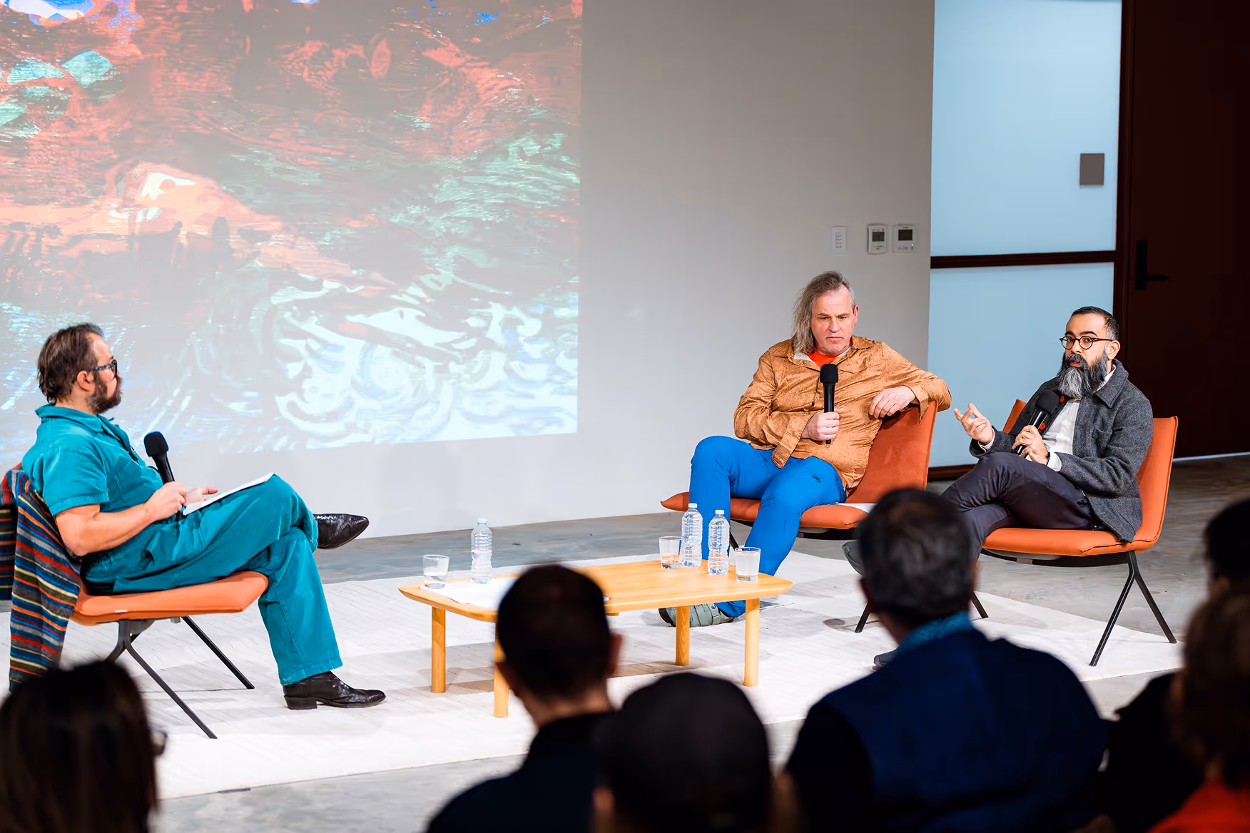 Three men in a panel discussion seated on orange chairs, holding microphones, with a wooden table and water bottles in front and an abstract colorful projection on the wall behind.