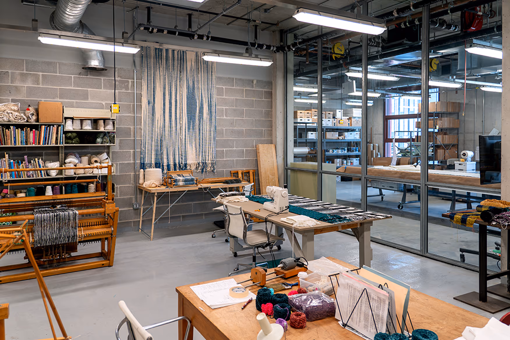 Art studio with weaving equipment, yarn, shelving with craft supplies, and a sewing machine on tables in a bright industrial space.
