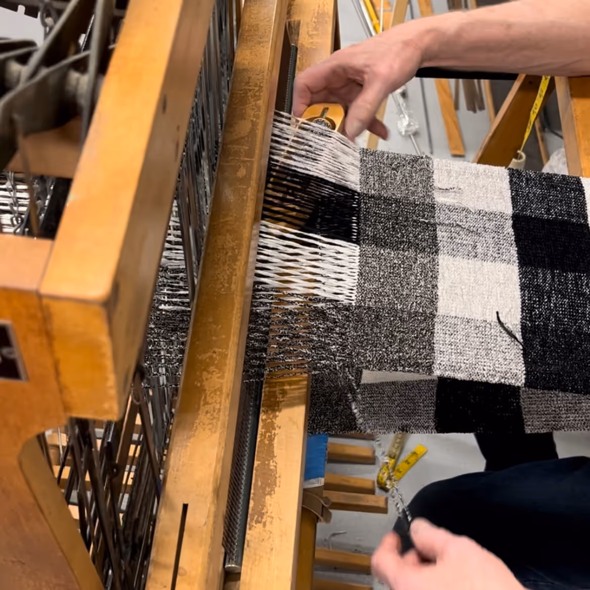Hands weaving a black and white checkered fabric on a wooden loom.