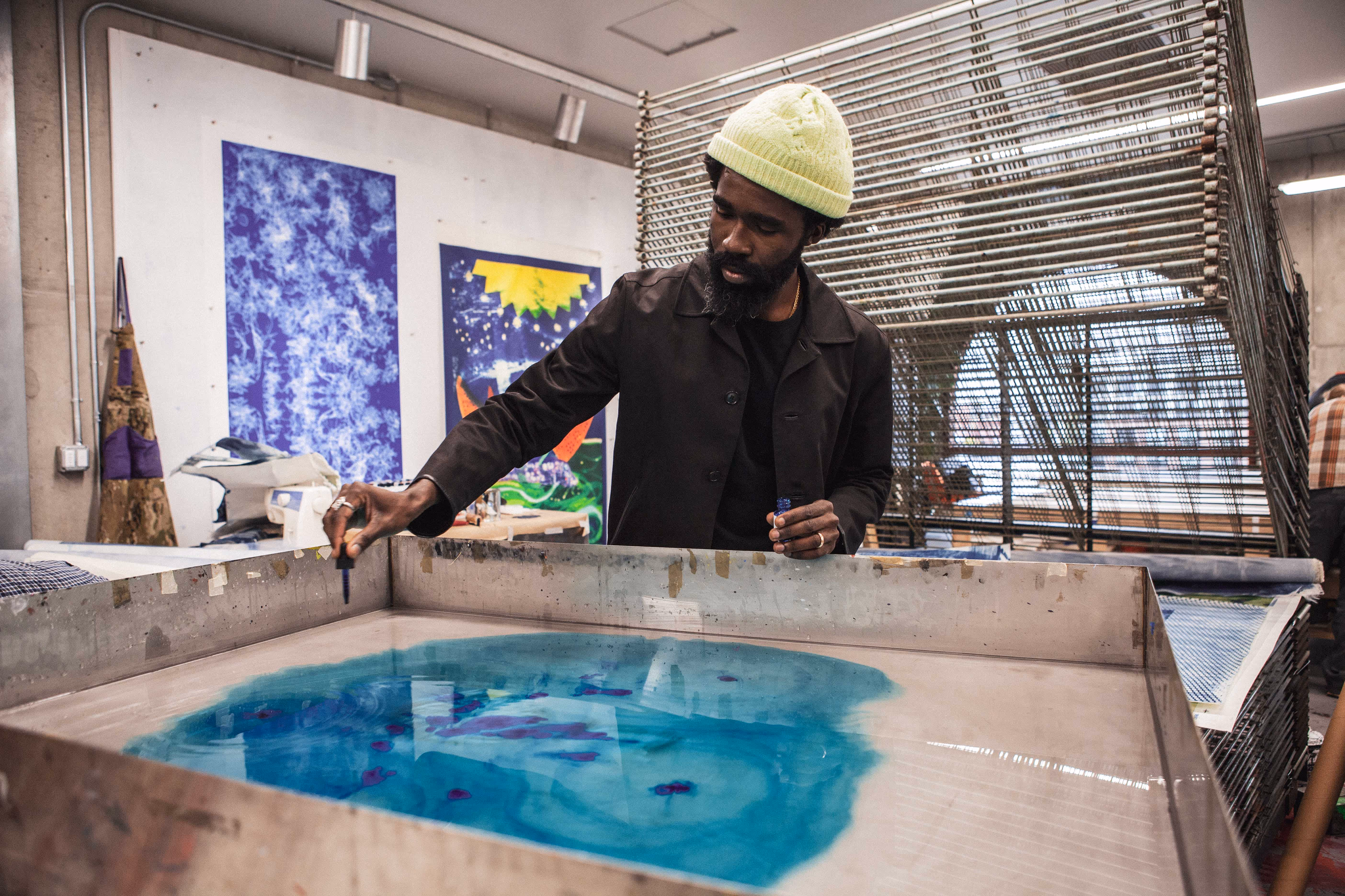 Man wearing a yellow beanie and dark jacket working with blue ink on a large flat surface in an art studio.
