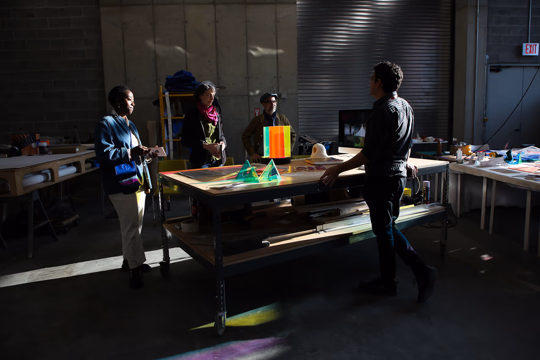 Four people standing around a workshop table with colorful translucent geometric shapes and materials in a concrete industrial space.