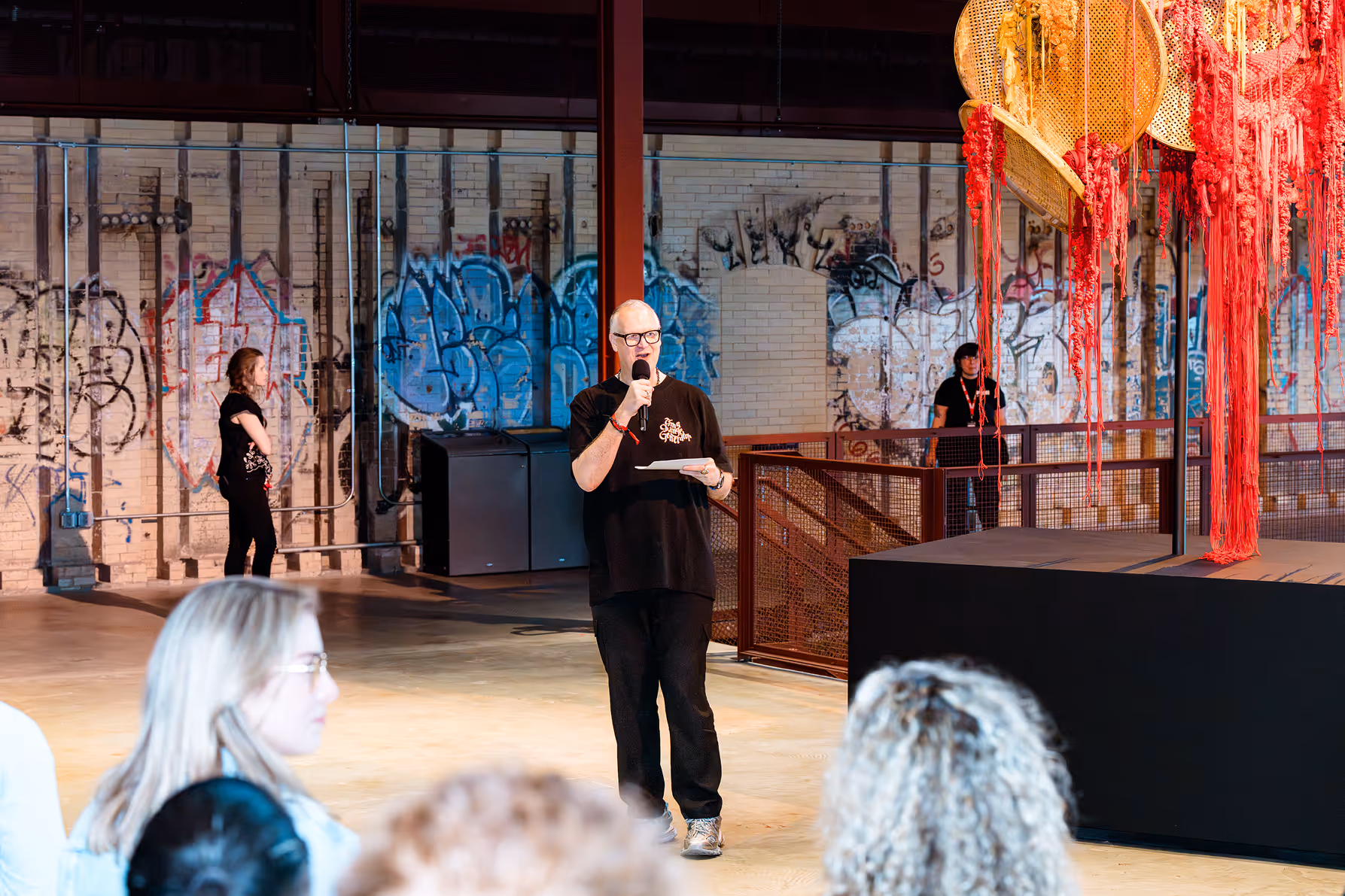 Man in black shirt and glasses speaking into a microphone in an industrial-style room with graffiti and hanging red decorations.