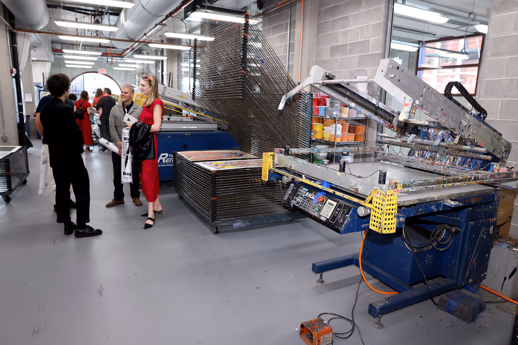 Group of people chatting inside a screen-printing workshop with printing machines and drying racks.