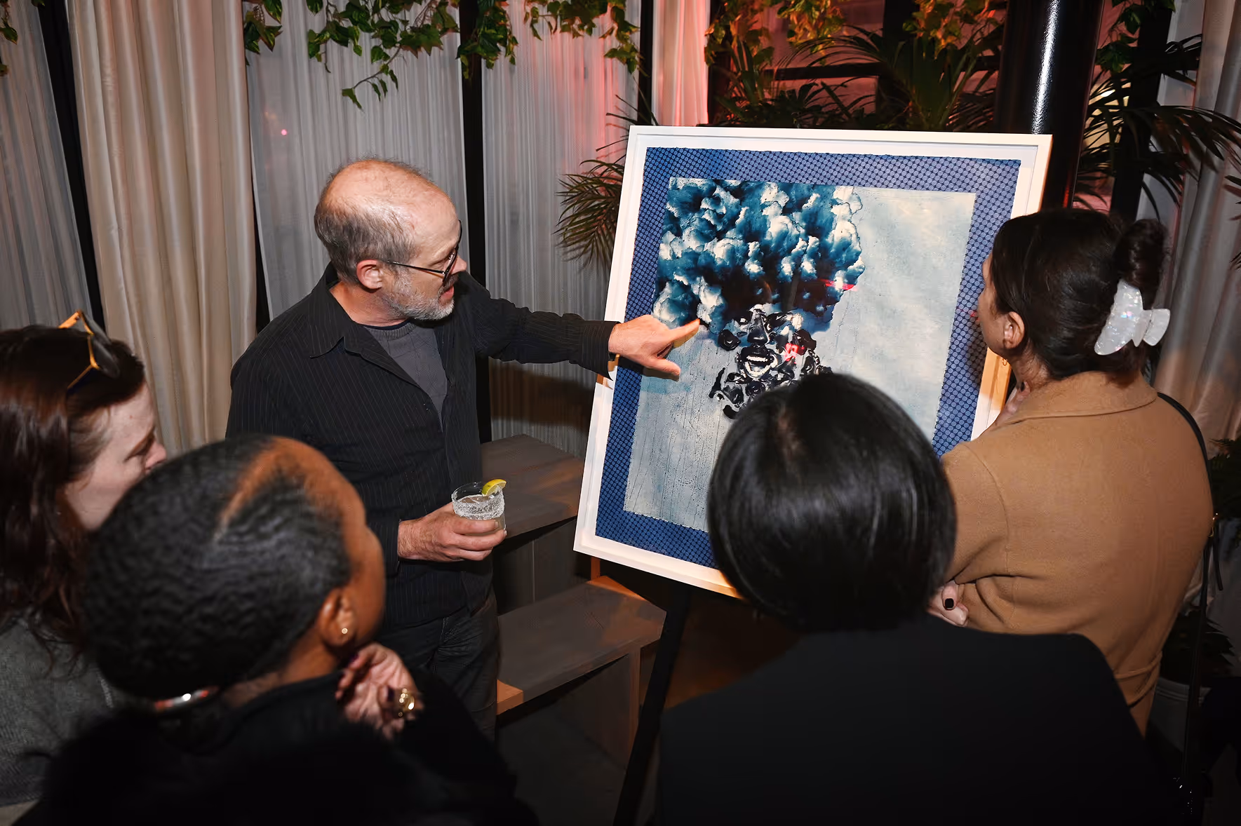 Man holding a drink and pointing to a framed abstract painting with blue and black tones while three women attentively look on.