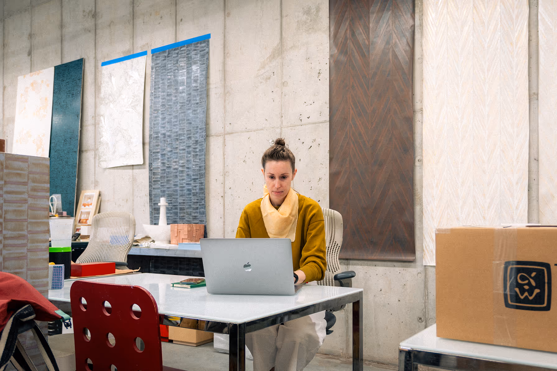 Woman in mustard sweater working on a laptop at a white table in an art studio with artwork on concrete walls.