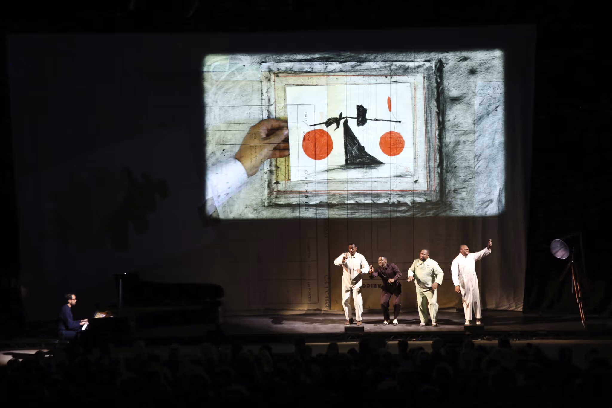 Four men performing on stage with a pianist on the left and a large projection of a hand holding a paper with a black and red abstract drawing.