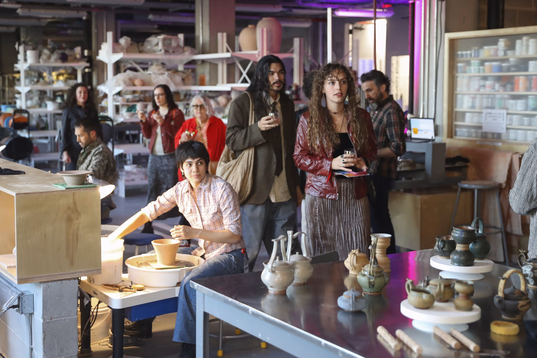 A group of people observing a pottery demonstration in a workshop, with ceramic pieces displayed on a table in the foreground.