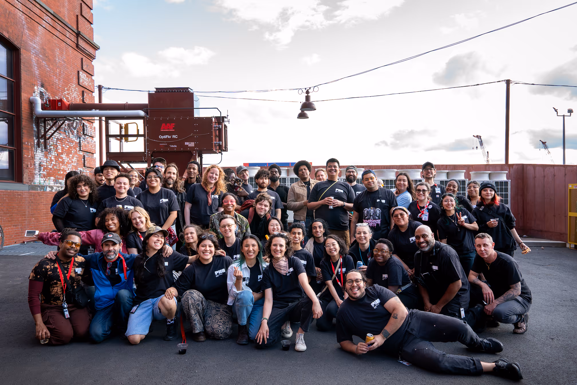 Large diverse group of people posing together outside near a brick building, many wearing black Powerhouse Arts t-shirts.