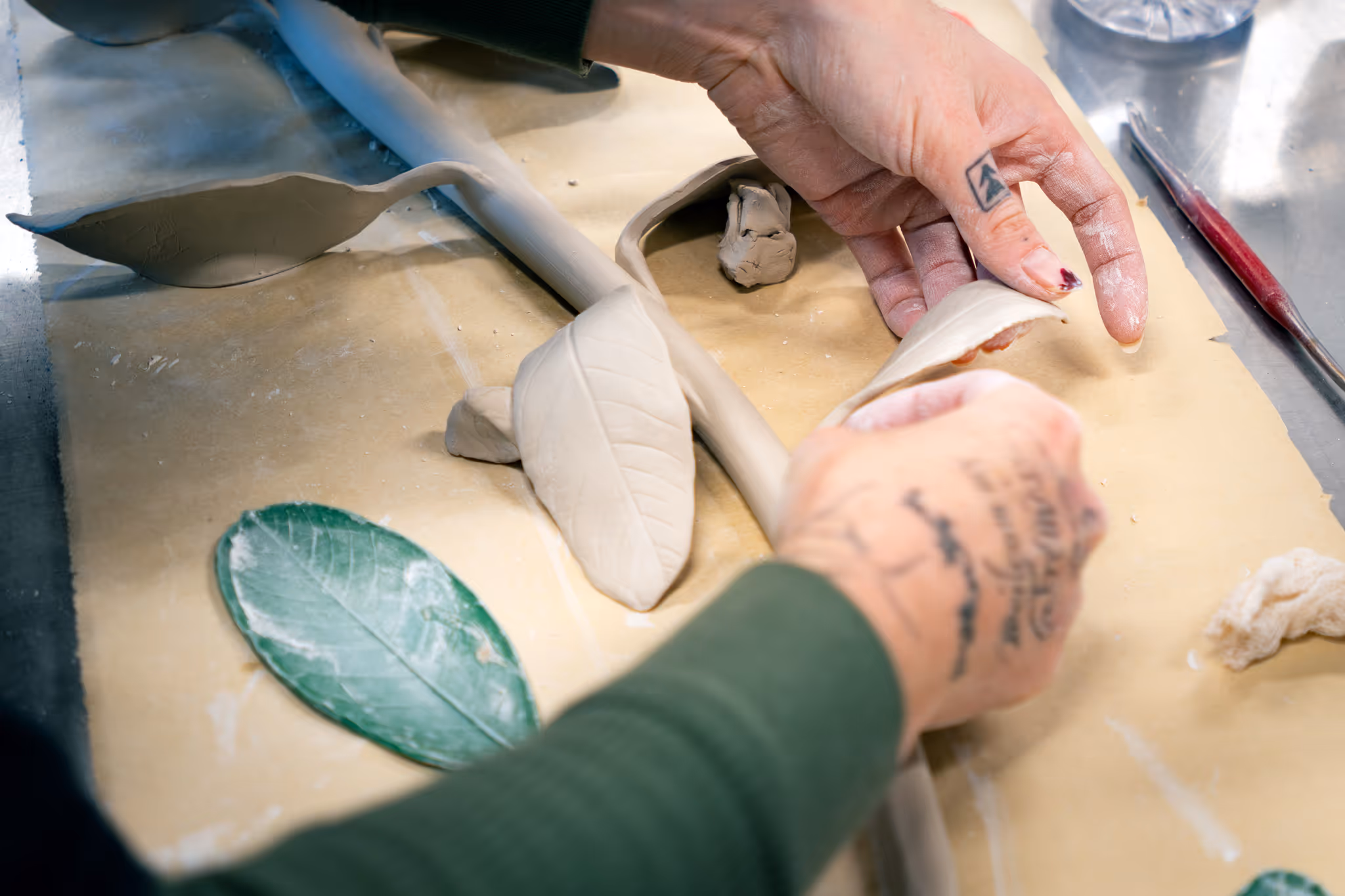 Hands shaping and assembling clay leaves and stem on a work surface for a ceramic sculpture.