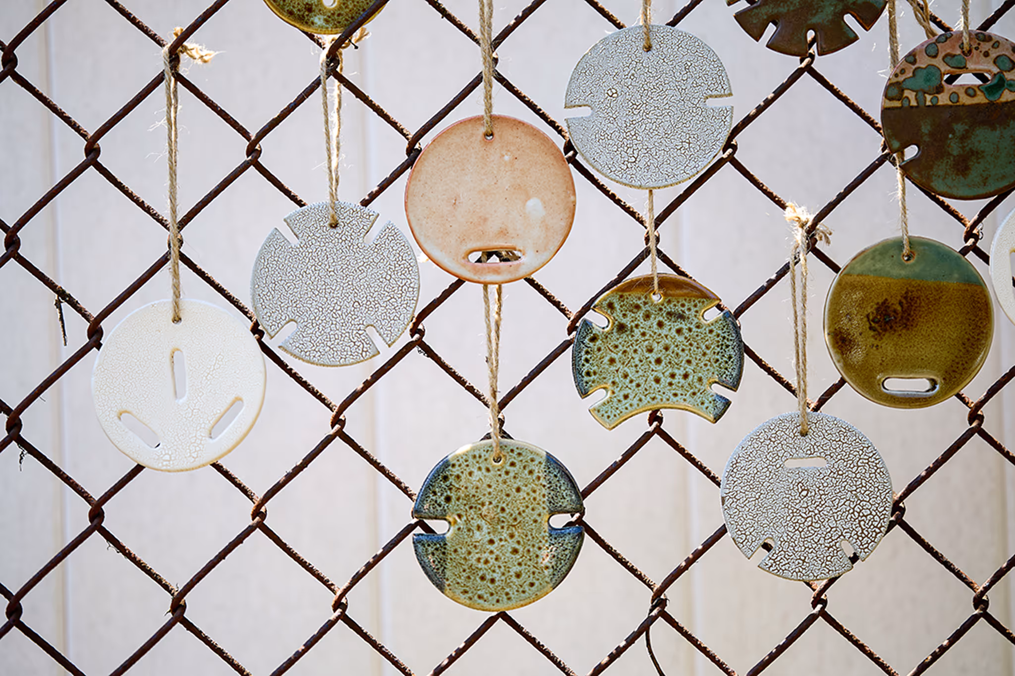 Various round ceramic ornaments with textured and glazed surfaces hanging from a rusty chain-link fence by twine.