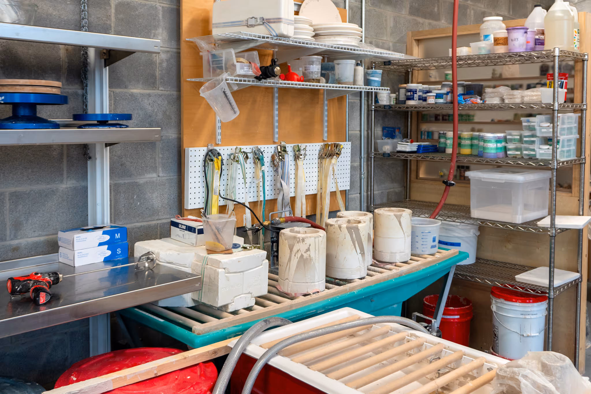 Workshop table with ceramic molds, measuring cups, rubber bands, and shelves stocked with containers and supplies for mold making and slip casting.