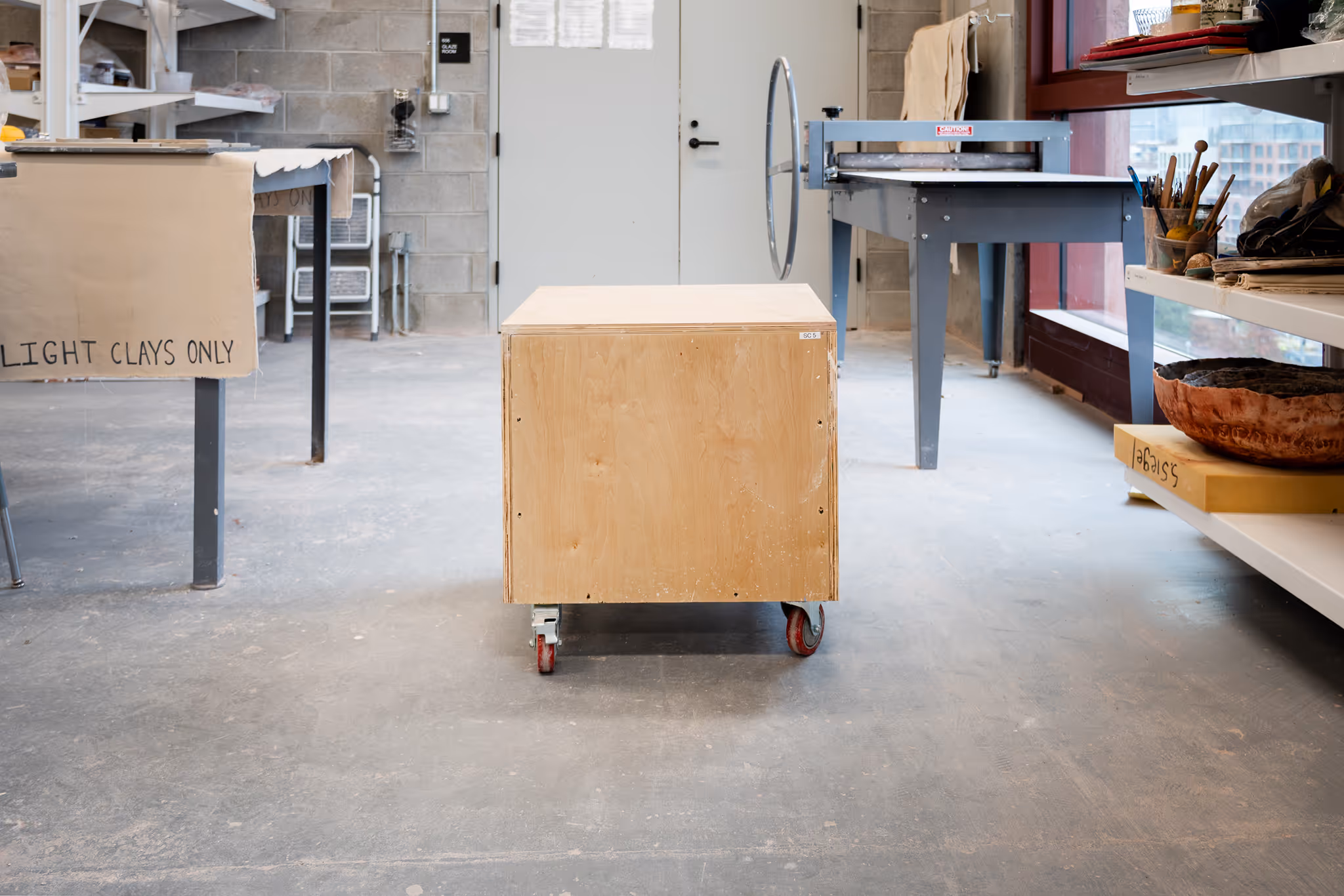 Wooden box on red caster wheels in a pottery studio with shelves, tools, and a pottery wheel.
