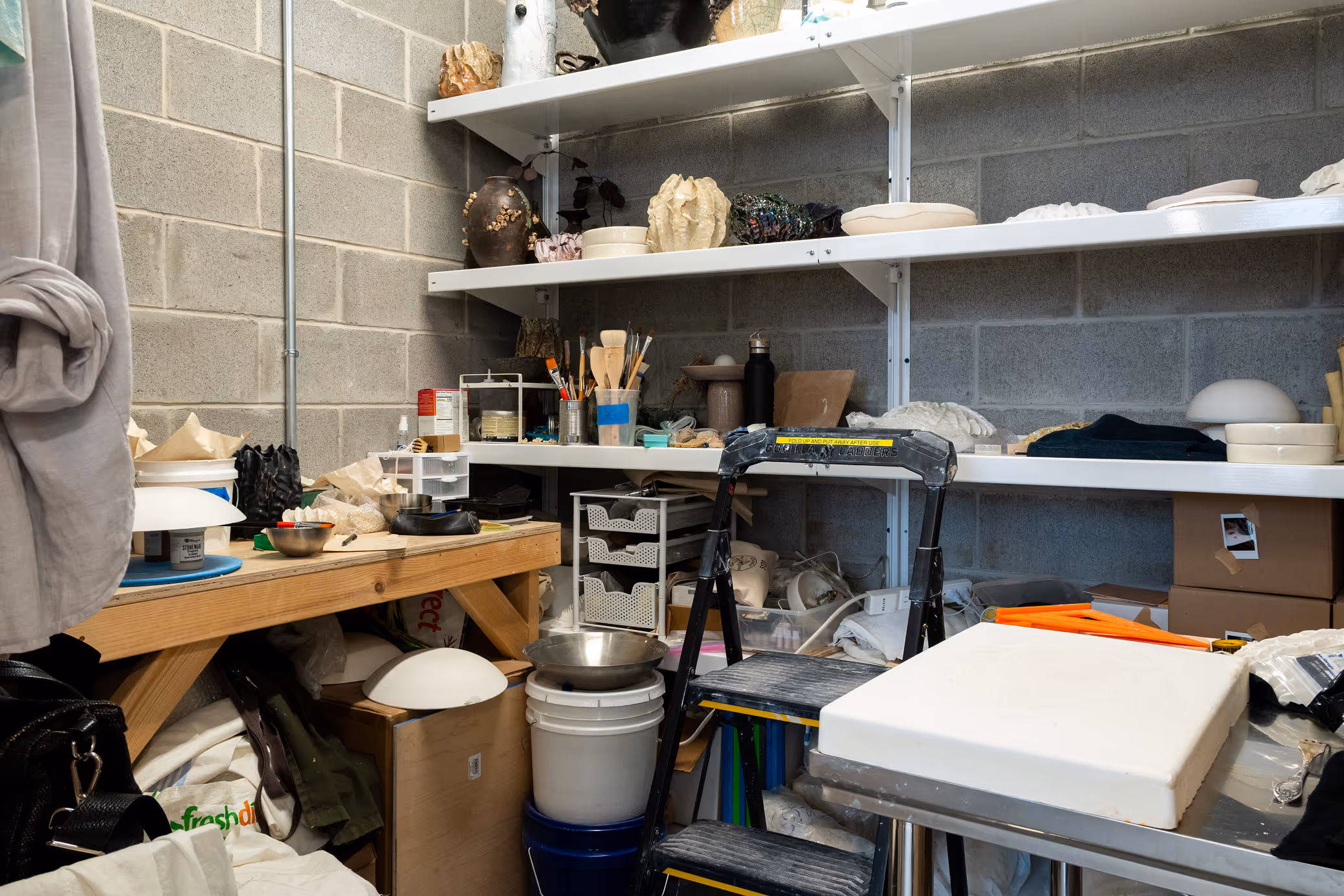 Cluttered workshop corner with wooden workbench, shelves holding ceramics and art supplies, a black step ladder, and various containers and tools.