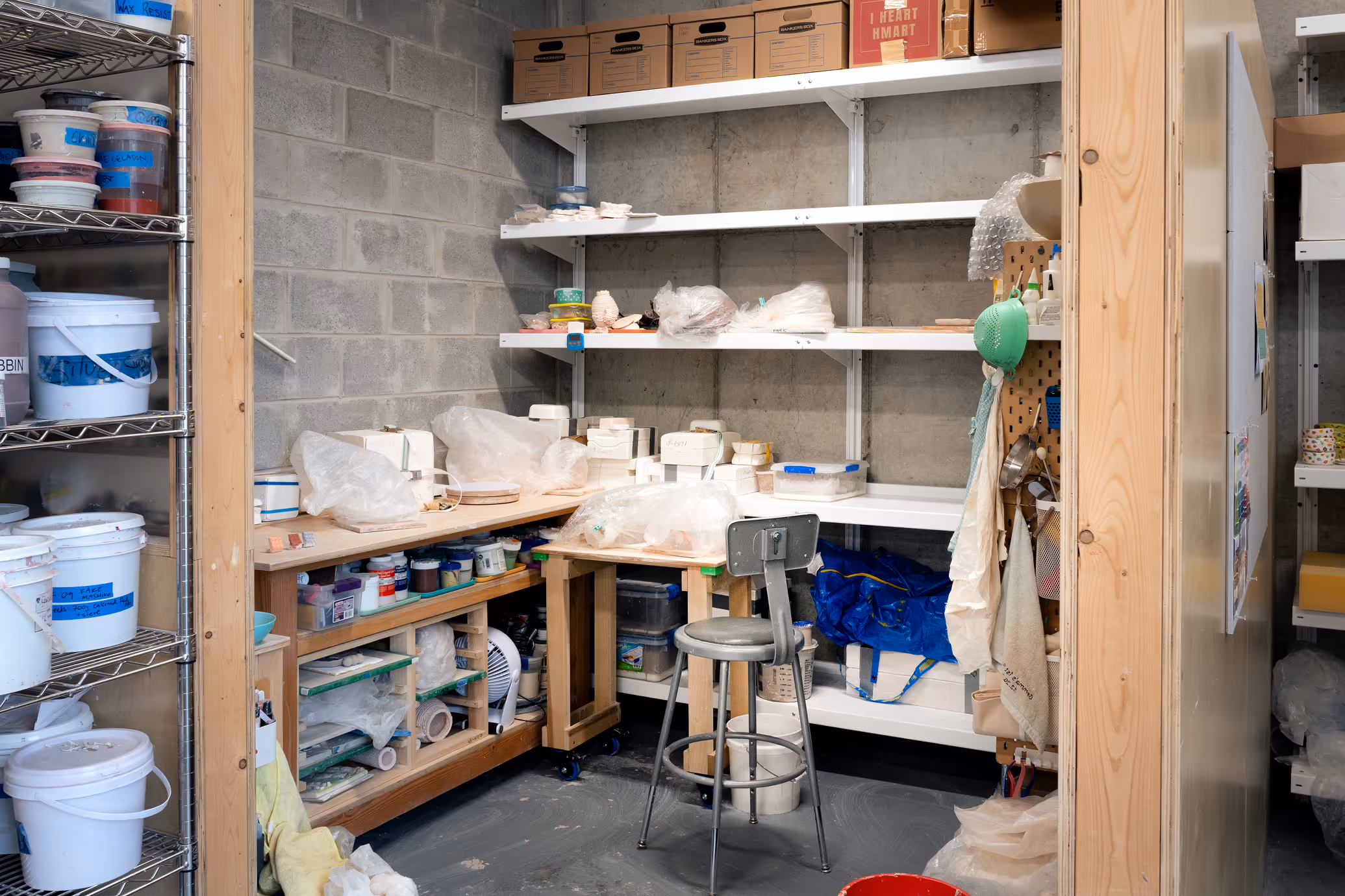 A storage room with shelves and counters holding containers, plastic-wrapped items, and various art supplies, with a metal stool in the center.