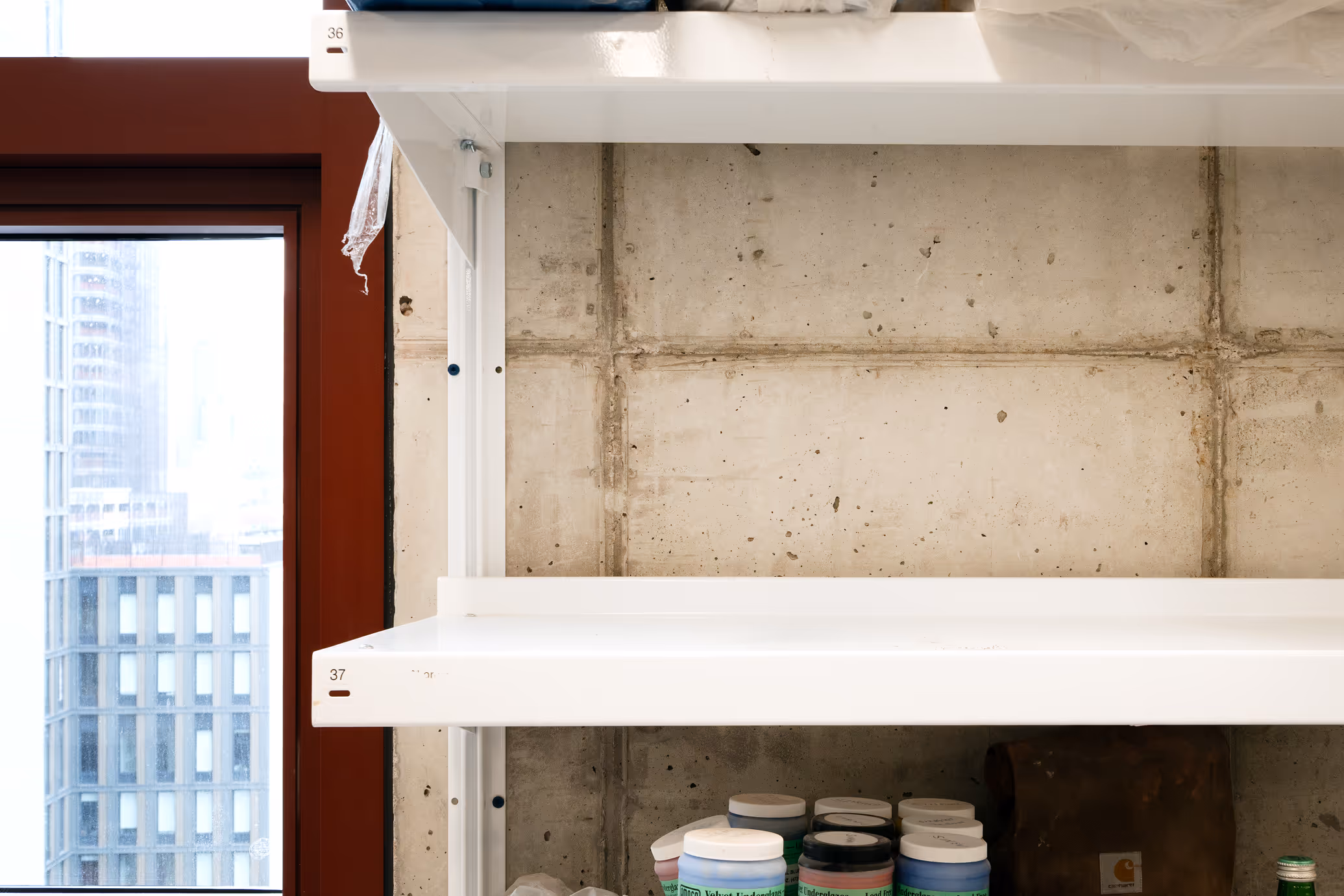 White metal shelves mounted on a concrete wall near a window with city buildings visible outside; lower shelf holds several small jars with colorful lids.